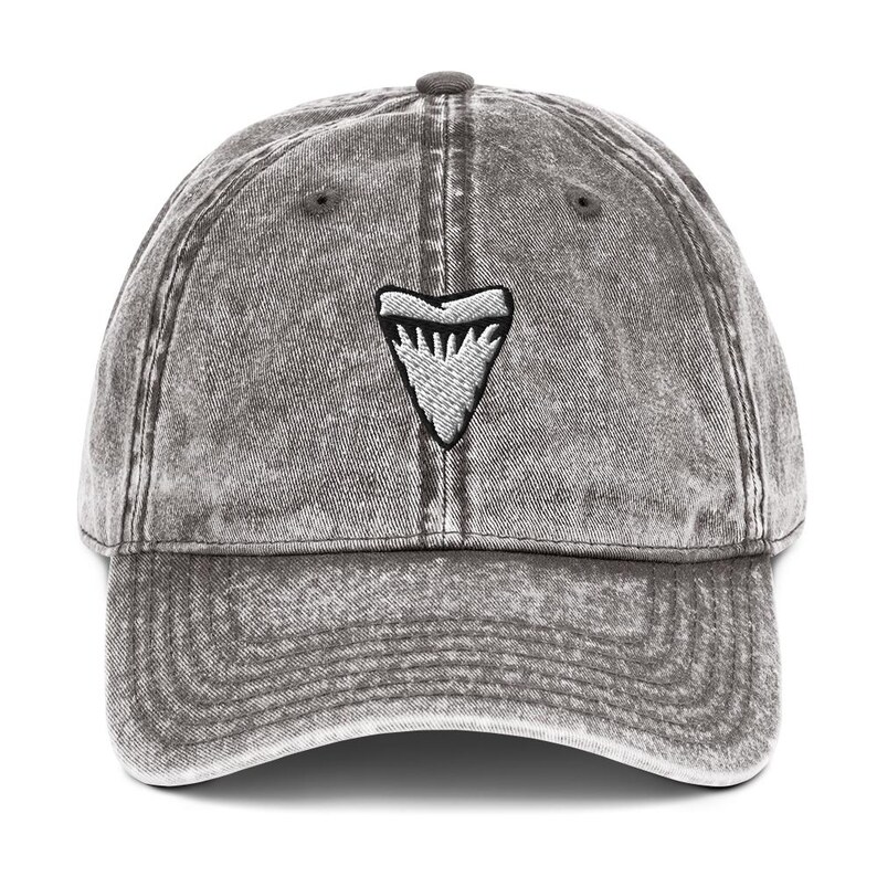 May include: A grey, distressed baseball cap featuring a white shark tooth emblem. The cap has a curved peak and a fabric strap. The hat is made of a soft, worn material, with a vintage look.