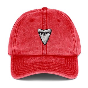 May include: Red baseball cap featuring a white and black shark tooth emblem. The cap has a curved peak and a faded, vintage appearance. It's a classic six-panel design, perfect for casual wear.