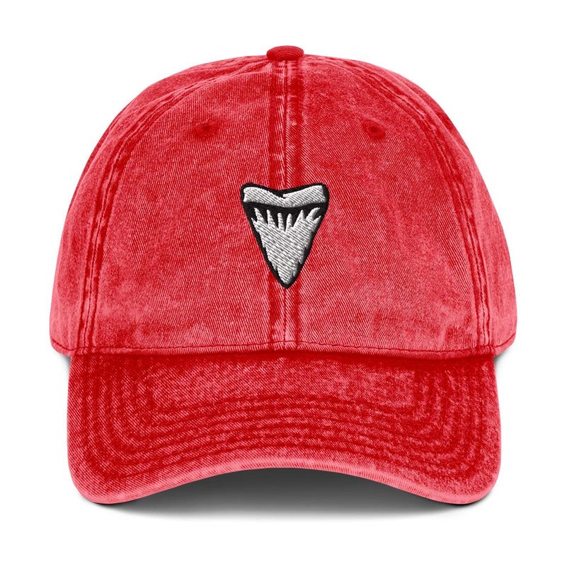 May include: Red baseball cap featuring a white and black shark tooth emblem. The cap has a curved peak and a faded, vintage appearance. It's a classic six-panel design, perfect for casual wear.