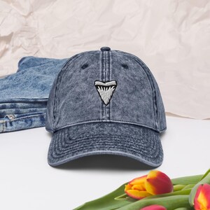 May include: A faded blue denim baseball cap with an embroidered shark tooth motif. The cap has a curved peak and a classic six-panel design. The shark tooth is white with grey detailing.