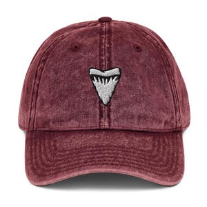 May include: A maroon baseball cap featuring a white and grey embroidered shark tooth emblem. The cap has a curved peak and a fabric strap. The hat is made of a soft, textured material.