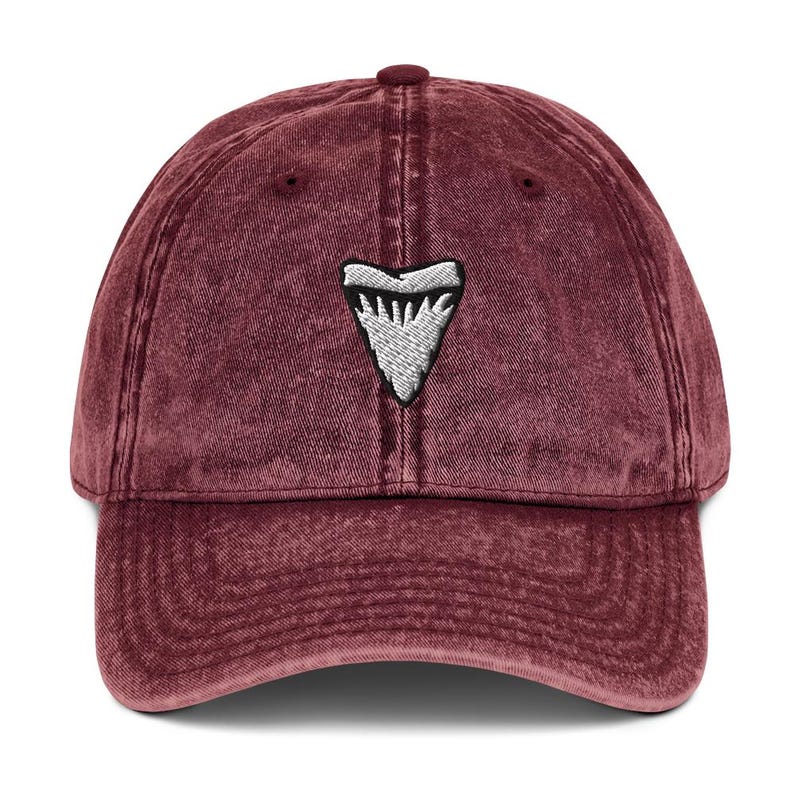 May include: A maroon baseball cap featuring a white and grey embroidered shark tooth emblem. The cap has a curved peak and a fabric strap. The hat is made of a soft, textured material.