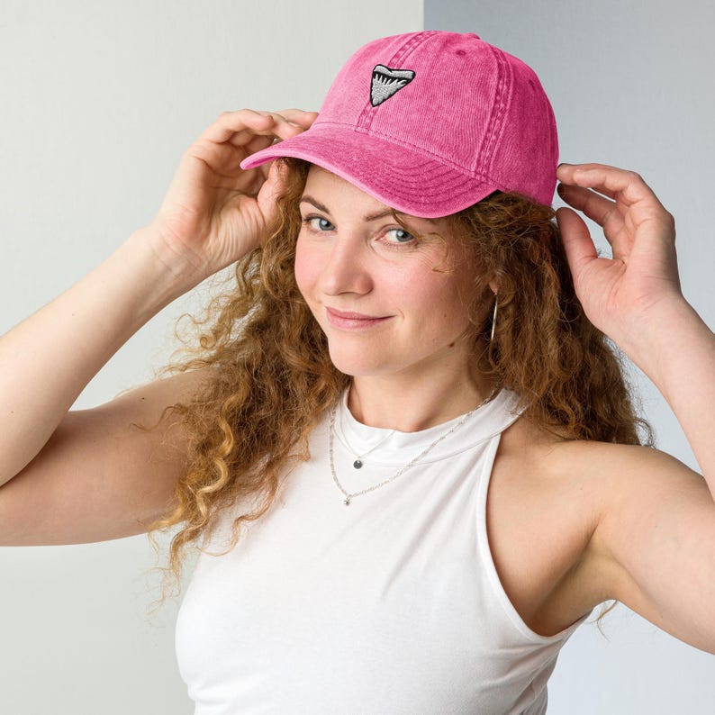 May include: A pink baseball cap featuring a shark tooth design. The cap is made of a soft, textured material and has a curved peak. The shark tooth design is embroidered in black and white.