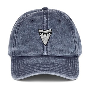 May include: A blue denim baseball cap with a distressed finish. The cap features a white embroidered shark tooth design on the front. The cap has a curved brim and an adjustable strap at the back.