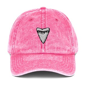 May include: A pink baseball cap with a faded effect. The cap has an embroidered shark tooth design in white and grey on the front. It features a curved peak and an adjustable strap at the back.