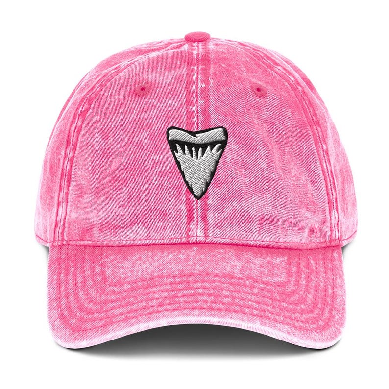 May include: A pink baseball cap with a faded effect. The cap has an embroidered shark tooth design in white and grey on the front. It features a curved peak and an adjustable strap at the back.