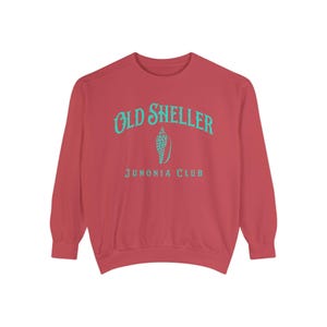 以下が含まれることがあります： 赤いスウェットシャツ。上部にターコイズ色の「OLD SHELLER」の文字がアーチ状に、下にターコイズ色の貝殻のグラフィック。貝殻のグラフィックの下には、ターコイズ色で「JUNONIA CLUB」の文字がプリントされています。