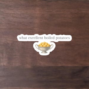 May include: A sticker showing a silver bowl filled with yellow boiled potatoes. Above the bowl is a white banner with the text "what excellent boiled potatoes" in a black, serif font. The sticker is on a dark brown wooden background.