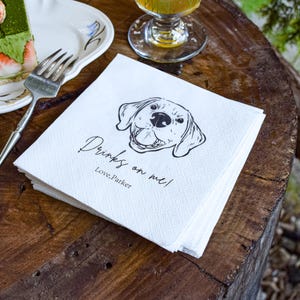 May include: A stack of white paper napkins with a black line drawing of a dog's face and the text "Drinks on me! Love, Parker" on a wooden table. A slice of cake with strawberries and a fork are on a plate.