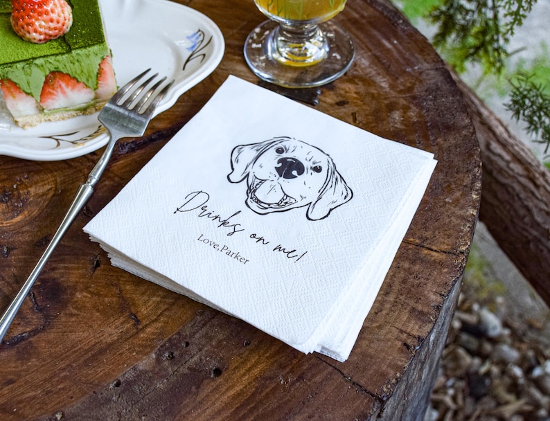 May include: A stack of white paper napkins with a black line drawing of a dog's face and the text "Drinks on me! Love, Parker" on a wooden table. A slice of cake with strawberries and a fork are on a plate.