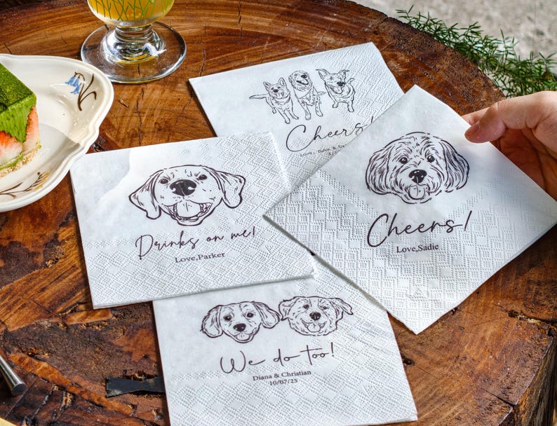 May include: White paper napkins with dog illustrations and text. One napkin says "Drinks on me! Love, Parker", another says "Cheers! Love, Sadie", and a third says "We do too! Diana & Christian 10/02/25".