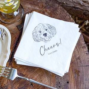 May include: A stack of white paper napkins with a black line drawing of a dog's face. The napkins have the word "Cheers!" and the text "Love, Sadie" printed on them. A glass of tea and a plate with cake and a fork are also visible.