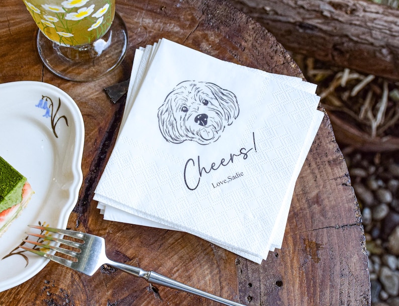 May include: A stack of white paper napkins with a black line drawing of a dog's face. The napkins have the word "Cheers!" and the text "Love, Sadie" printed on them. A glass of tea and a plate with cake and a fork are also visible.