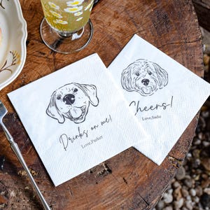 May include: Two white paper napkins with dog illustrations and text. One napkin reads "Drinks on me! Love Parker" and the other says "Cheers! Love, Sadie". The napkins are on a wooden surface.