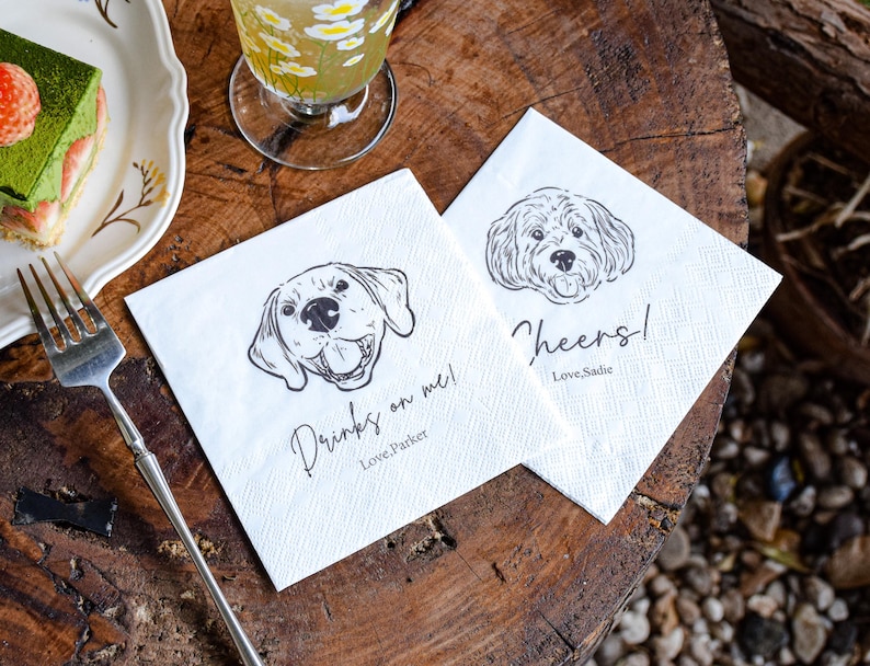 May include: Two white paper napkins with dog illustrations and text. One napkin reads "Drinks on me! Love Parker" and the other says "Cheers! Love, Sadie". The napkins are on a wooden surface.