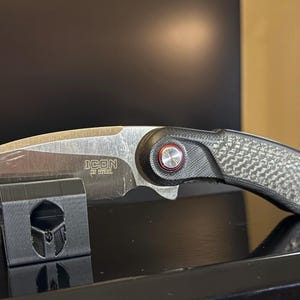 Könnte beinhalten: Ein silbernes und schwarzes Klappmesser mit dem Text "ICON by STEEL" auf der Klinge. Der Griff hat eine strukturierte Kohlefaseroptik. Das Messer liegt auf einem schwarzen Ständer.