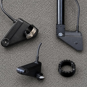 May include: A collection of black electronic components, including a sensor with a hose clamp, a bracket, and a circular gear-like piece. The components are arranged on a gray textured surface, suggesting a technical or mechanical theme.