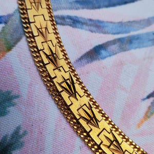 May include: A gold chain necklace featuring a textured, geometric design. The chain is made up of interlocking links with a repeating pattern of squares and triangles.