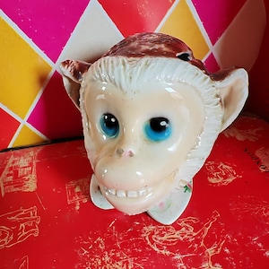 May include: A ceramic monkey head planter with blue eyes and a toothy grin. The planter is white with brown fur on top and has a floral design around the neck.