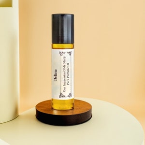 Op de afbeelding: Een doorzichtige glazen fles met een zwarte dop en een wit etiket met de tekst "Delina Our Impression Of de Marly Pure Perfume Oil". De fles staat op een rond houten platform.