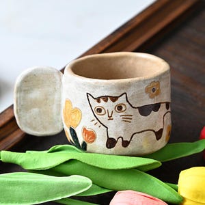 Peut inclure: Une tasse en céramique blanche avec un motif de chat et de fleurs. La tasse a une anse en forme d'oreille de chat.
