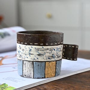 Pode incluir: Uma caneca de cerâmica feita à mão com um design listrado em marrom, branco, azul e amarelo. A caneca tem uma alça marrom costurada.