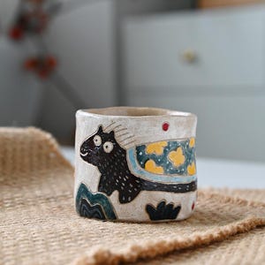 May include: A white ceramic mug with a black cat design. The cat has large eyes and is wearing a blue and yellow patterned blanket. The mug is hand-painted and has a rustic look.