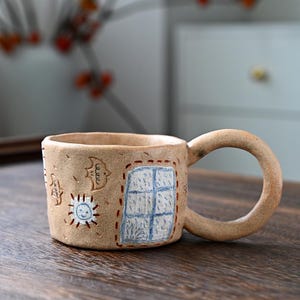 Peut inclure: Une tasse en céramique beige avec un motif peint à la main d'une fenêtre, d'un soleil et d'un oiseau. La tasse a une poignée arrondie et une surface texturée.