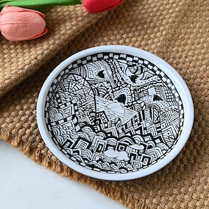 Peut inclure: Une assiette en céramique blanche avec un motif abstrait noir et blanc. Le motif présente une variété de formes géométriques et de motifs, y compris des cercles, des carrés et des lignes.