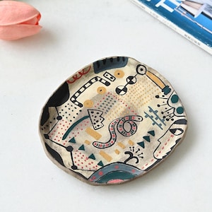 Puede incluir: Plato de cerámica pintado a mano con un diseño abstracto. El plato presenta una base de color crema con detalles en negro, rojo, amarillo y turquesa. La forma irregular y la obra de arte única lo convierten en una pieza decorativa.