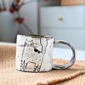 Könnte beinhalten: Eine weiße Keramiktasse mit einer schwarz-weißen Illustration einer Katze. Die Tasse hat eine gesprenkelte Glasur und einen abgerundeten Griff.