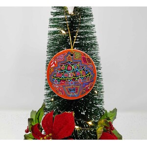Può includere: Un addobbo natalizio rotondo di colore arancione con un design geometrico colorato. L'addobbo è appeso a un albero di Natale artificiale verde.