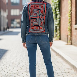 May include: A denim jacket with a colorful embroidered design on the back. The embroidery features geometric shapes in red, teal, and orange. The jacket is paired with blue jeans and white sneakers.