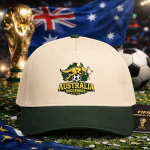 Puede incluir: Gorra de béisbol crema y verde oscuro con el texto "Australia Socceroos" y un logotipo de canguro. La gorra está colocada frente a un balón de fútbol, la bandera australiana y un trofeo.