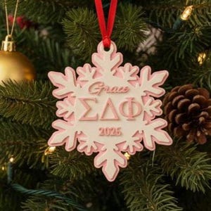 May include: A snowflake-shaped Christmas ornament with a pink and white design hangs on a decorated Christmas tree. The ornament has the name "Grace", Greek letters, and the year "2025" engraved on it. The tree is adorned with red, gold, and silver ornaments.