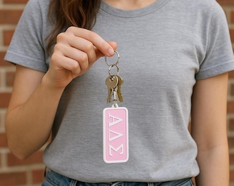May include: A rectangular pink keychain with a white border featuring the Greek letters Alpha, Delta, and Sigma in white. The keychain is attached to a silver metal split ring holding two silver keys, showcasing a clean and simple design.