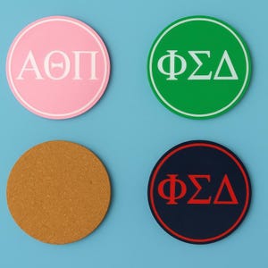 May include: Four round coasters on a blue background. One pink coaster with white Greek letters, one green coaster with white Greek letters, one cork coaster, and one navy blue coaster with red Greek letters.