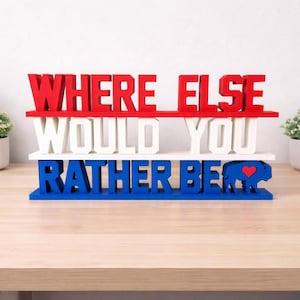 May include: A three-tiered wooden sign with the text "WHERE ELSE WOULD YOU RATHER BE" in red, white, and blue. The bottom tier features a blue buffalo with a red heart. A decorative item for home or office.