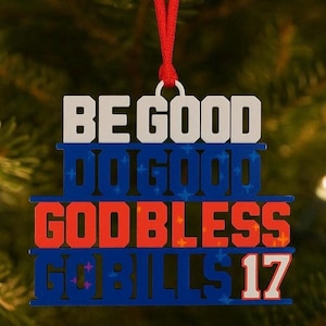 May include: A layered Christmas ornament with the words "BE GOOD," "DO GOOD," "GODBLESS," and "BILLS 17" in white, blue, and red. The ornament is suspended by a red string against a blurred green background.