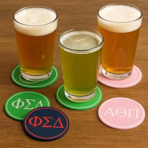 May include: Three glasses of beer with foam, resting on coasters. The coasters are green, pink, and navy blue, with Greek letters. The beer is light amber and green. The coasters are round.