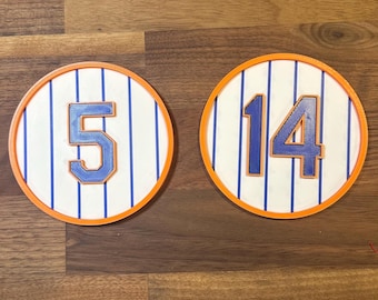 May include: Two round, orange-rimmed coasters with white and blue striped designs. The coasters feature the numbers "5" and "14" in blue.