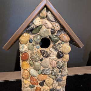 May include: A decorative birdhouse with a unique stone mosaic exterior. The house features a variety of colorful stones in shades of brown, grey, yellow, and green. The roof is made of brown wood, and there is a circular entrance hole.