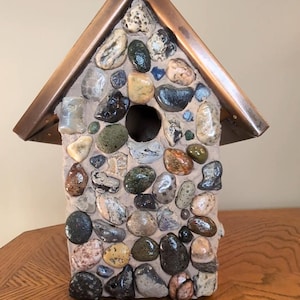 May include: A handcrafted birdhouse featuring a copper roof and a body adorned with a mosaic of multi-coloured stones. The stones vary in size and hue, including brown, grey, green, and black. It has a circular entrance.