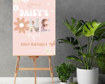May include: Pink and brown floral first birthday sign with the text "Welcome to Daisy's One derful First Birthday 28.06.2023". The sign has a photo of a baby girl in the letter "O".