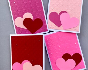 May include: Four handmade Valentine's Day cards with pink and red hearts on a white background. Each card has a different pattern of small hearts.