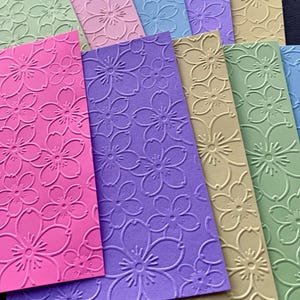 May include: Assortment of embossed cardstock paper in various colors, including pink, purple, green, and blue. Each sheet features a repeating pattern of embossed cherry blossoms.