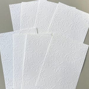 May include: A stack of white paper cards with an embossed maple leaf pattern. The cards are arranged in a fan shape, showcasing the detailed texture of the leaves. The background is a neutral gray.