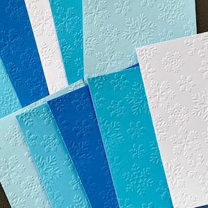 May include: A stack of blue and white cardstock paper with an embossed snowflake pattern. The paper is arranged in shades of blue, from light to dark.