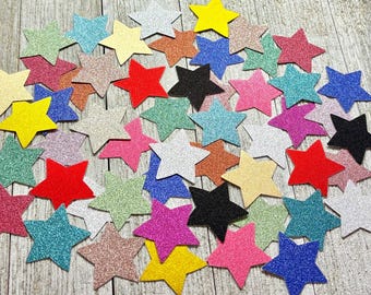 May include: A collection of glittery star-shaped embellishments in various colors, including red, yellow, blue, pink, green, and black. These decorative stars are scattered across a wooden surface, creating a festive and eye-catching display.
