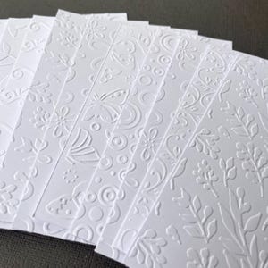 May include: A stack of white embossed paper cards with various floral and geometric designs. The cards are perfect for crafting, scrapbooking, or card making.